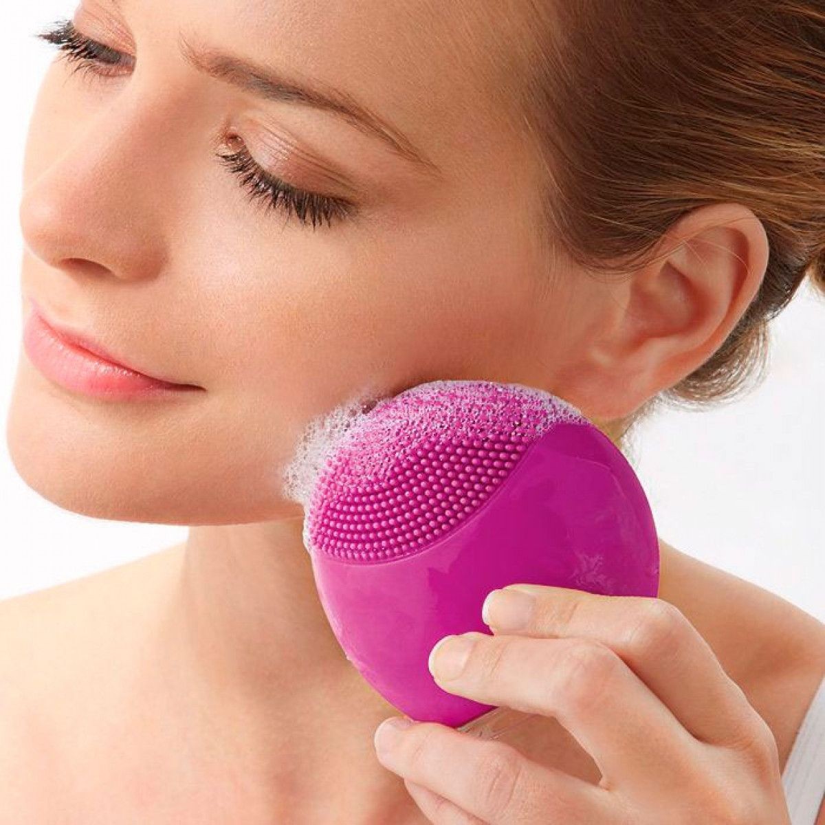 Person using a pink facial cleansing brush on their cheek near the ear, close-up view.