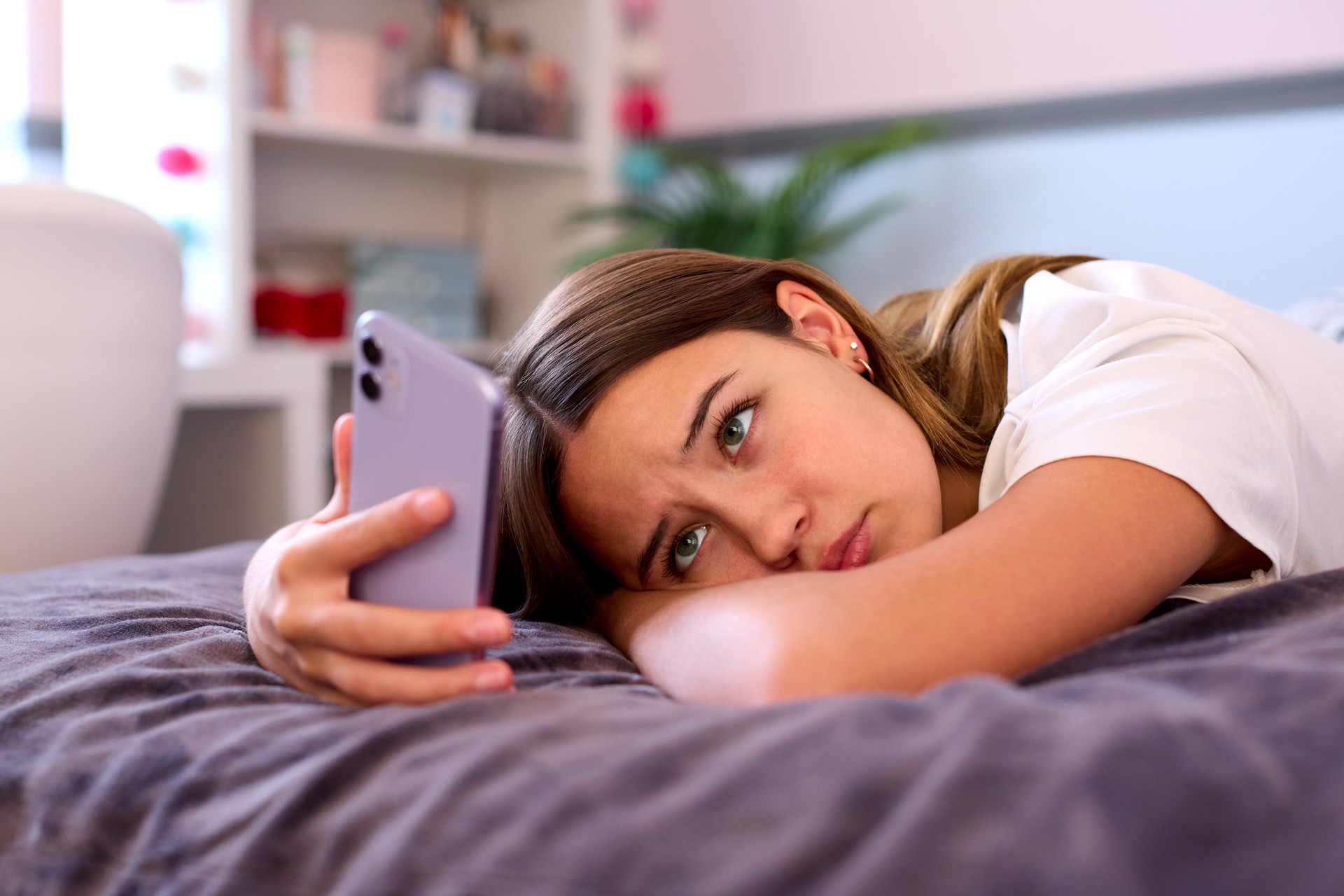 Depressed Teenage Girl Lying On Bed At Home Looking At Mobile Phone