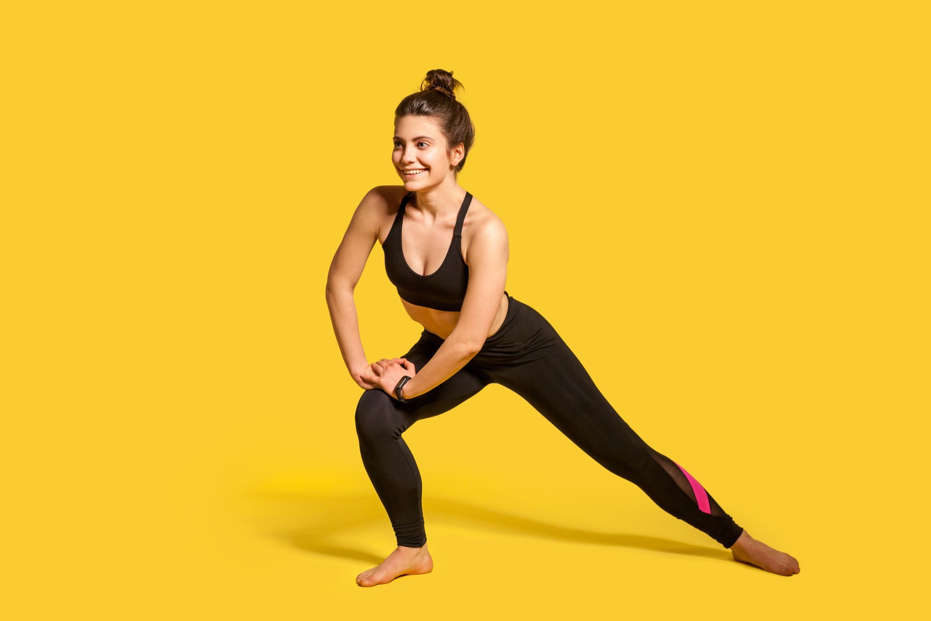 Happy athletic girl with hair bun in tight sportswear doing lower body sport exercise, stretching legs