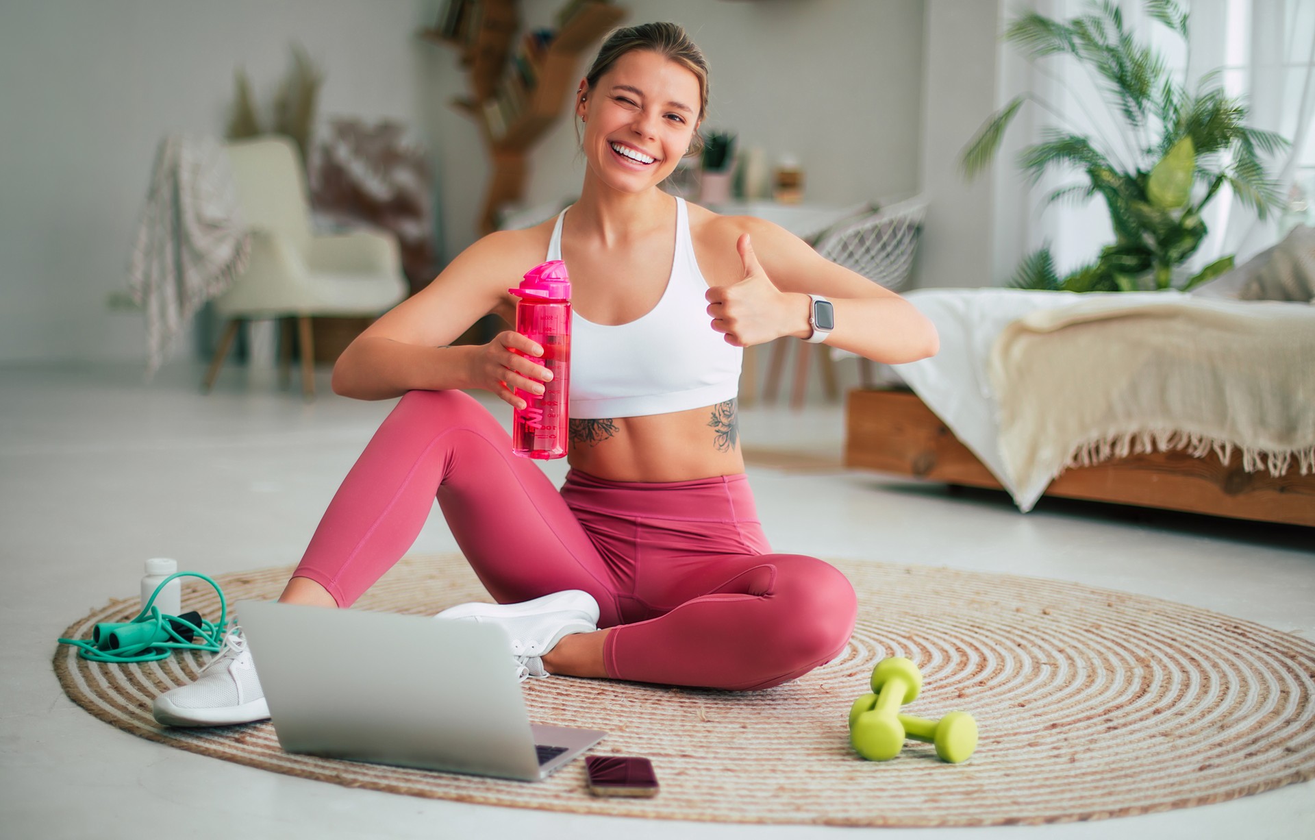 Sports trainer or teacher of Pilates online. Happy blond female in sportswear sitting on the floor with laptop after exercises and showing her thumb up on camera with a smile.