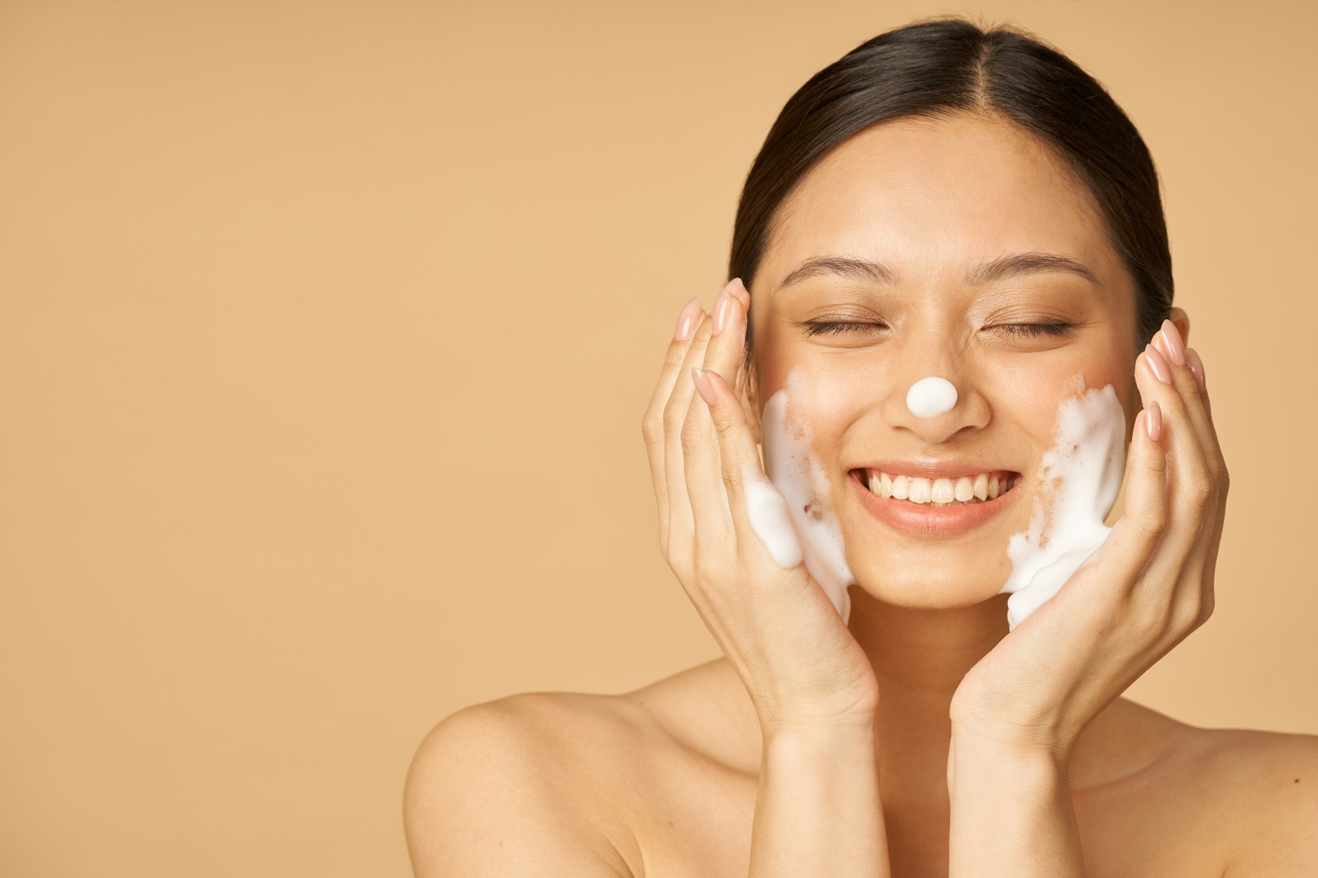 Studio portrait of pleased young woman smiling with eyes closed while applying gentle foam facial cleanser isolated over beige background