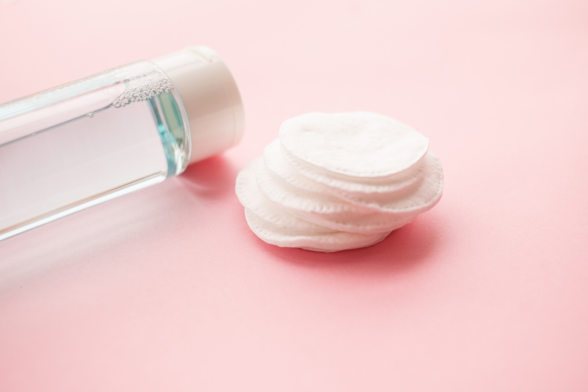 bottle with tonic or micellar cleansing water with fresh herbs and flowers and cotton pads on pink background, top view. Beauty, skin, hair or body care concept. selective focus