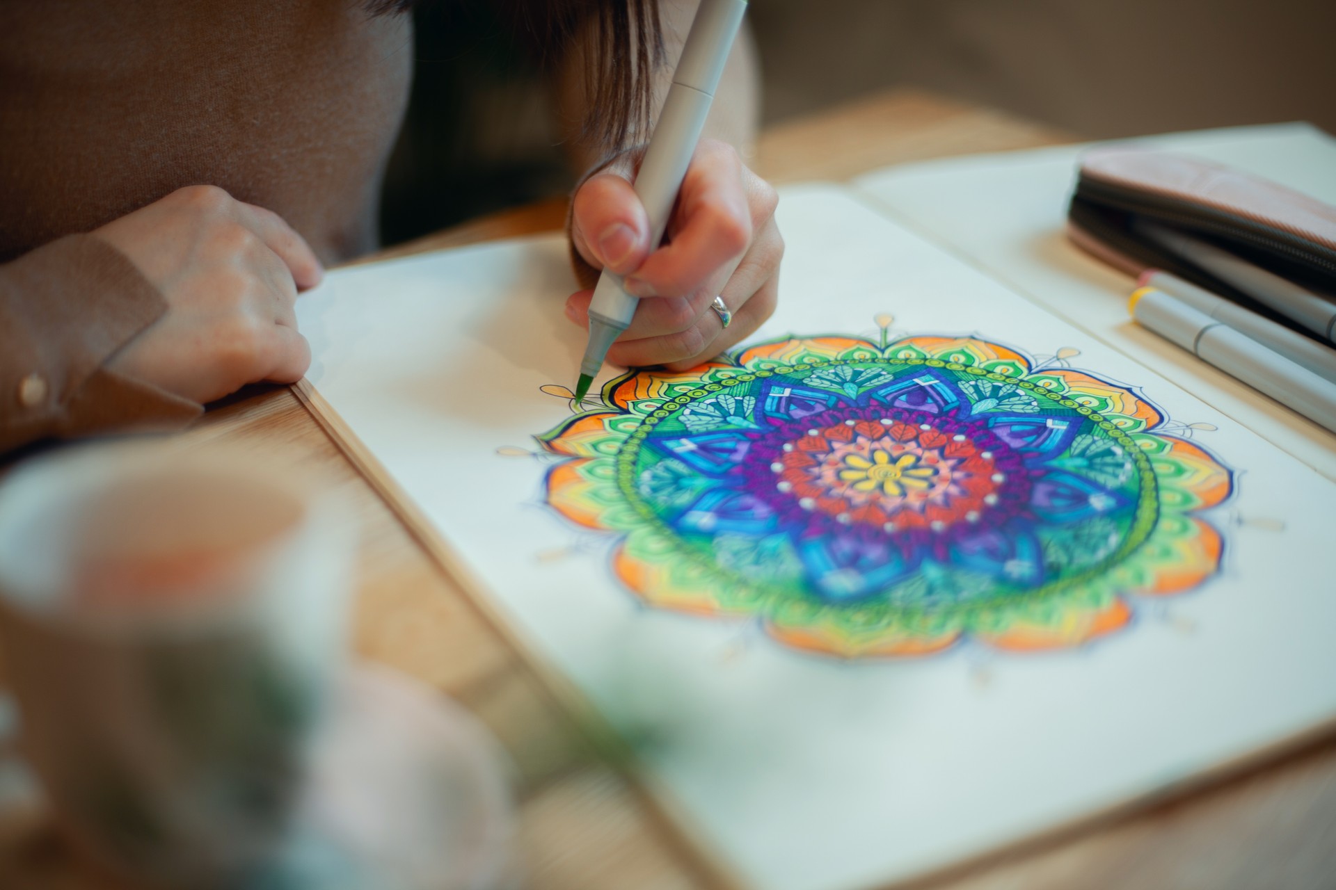 Young woman colouring mandala with markers and white rosary on table with cup of coffee at home